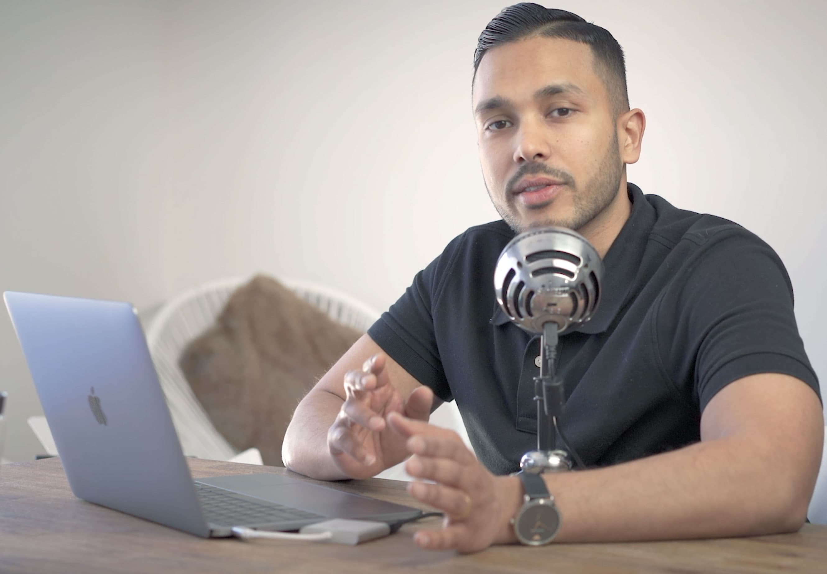 A man speaking into a silver microphone while gesturing at a desk with a laptop.